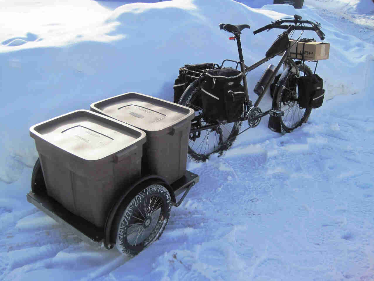Right side view of a Surly Troll bike with a loaded bike trailer hitched on back, parked against a high snow bank